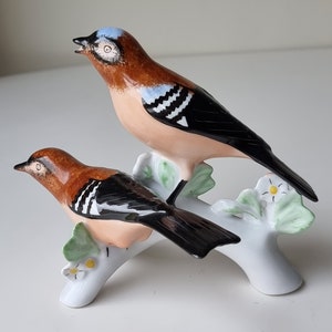 Puede incluir: Una figurita de porcelana de dos pájaros marrones y negros posados en una rama blanca con hojas verdes y flores amarillas.