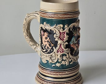 May include: A ceramic beer stein with a green and white design. The stein features a raised relief of a cherub playing a trumpet and a couple in traditional clothing. The stein has a handle and a wide base.