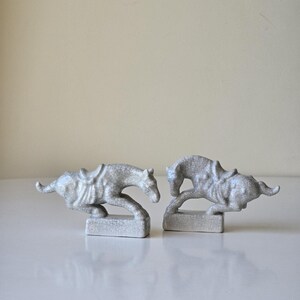 May include: Two white ceramic horse figurines with saddles, each standing on a small base. The horses are in a running pose, one with its head down and the other with its head up.