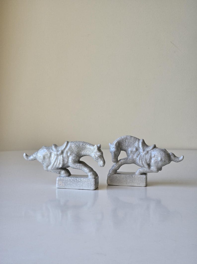 May include: Two white ceramic horse figurines with saddles, each standing on a small base. The horses are in a running pose, one with its head down and the other with its head up.