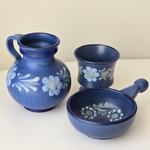 Könnte beinhalten: Ein Set aus drei blauen Keramikstücken: ein Krug, eine kleine Tasse und eine Schale mit Henkel. Jedes Stück ist mit einem Blumenmuster in Weiß und Hellblau verziert. Der Krug hat einen Henkel und einen runden Körper.