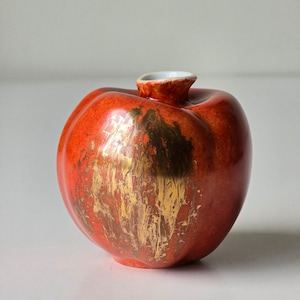 May include: A red ceramic vase shaped like an apple with gold accents. The vase has a smooth, glossy finish.