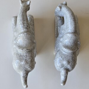 May include: Two white ceramic elephant figurines with a crackled glaze finish. The elephants are facing opposite directions and are in a seated position.