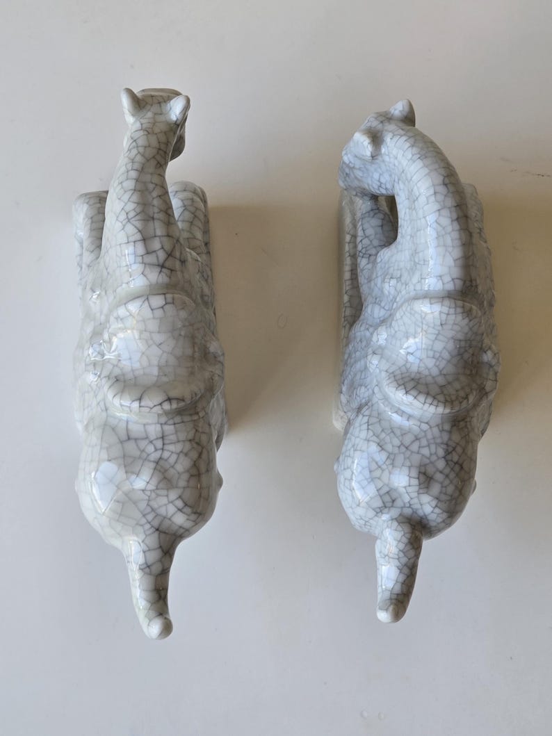 May include: Two white ceramic elephant figurines with a crackled glaze finish. The elephants are facing opposite directions and are in a seated position.