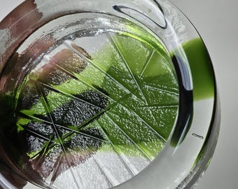 May include: A clear glass ashtray with a green, geometric design. The ashtray has a raised rim and a slightly indented center.