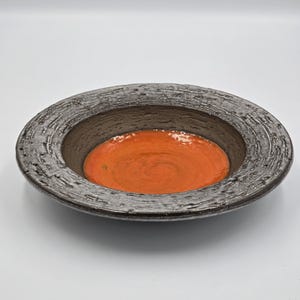 May include: A ceramic bowl with a textured, dark grey rim and a bright orange interior. The bowl has a circular shape with a wide rim, suitable for serving or display. The interior has a glossy finish. The bowl's diameter is approximately 25 cm.