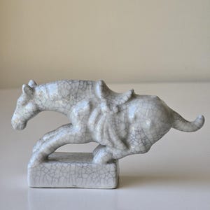 May include: A white ceramic horse figurine with a cracked glaze finish. The horse is depicted in a running pose with a saddle on its back. The figurine is standing on a small square base.