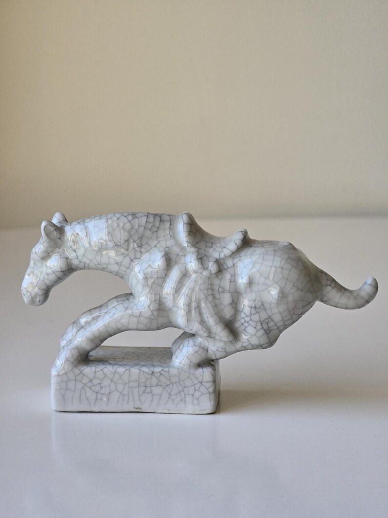 May include: A white ceramic horse figurine with a cracked glaze finish. The horse is depicted in a running pose with a saddle on its back. The figurine is standing on a small square base.