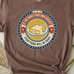 以下が含まれることがあります： 黄色い背景の円形グラフィックが特徴の茶色の半袖Tシャツ。デザインには皿に乗ったグレービーソースのかかったビスケットが描かれており、白い星が並ぶ赤と青の縁取りと、IF YOU MIND YOUR OWN BISCUIT EVERYTHING WILL BE GRAVYというテキストが添えられています。