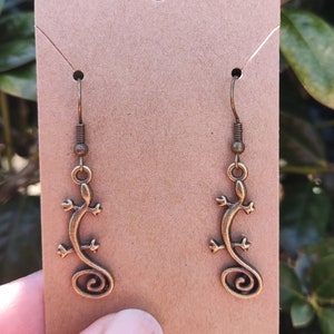 May include: A pair of bronze-colored earrings featuring a stylized lizard design. The earrings are hanging from a brown card.