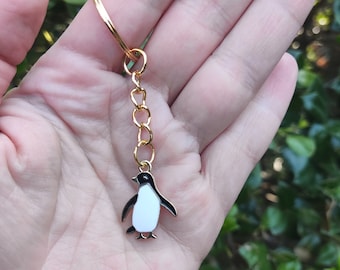 May include: A gold keychain with a black and white enamel penguin charm. The penguin is facing forward with its wings outstretched.