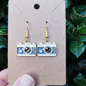 Puede incluir: Un par de pendientes de cámara dorados con detalles en esmalte azul y blanco. Los pendientes están en una tarjeta marrón.