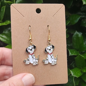 May include: A pair of gold dangle earrings featuring a cartoon Dalmatian puppy with black spots on a white background. The puppy is wearing a red collar.