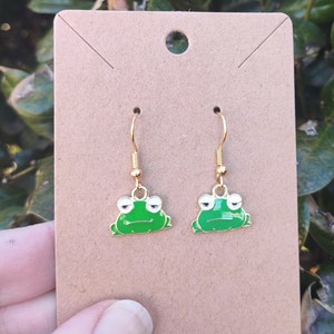 May include: A pair of gold dangle earrings with green frog charms. The frogs have white eyes and a slightly sad expression.