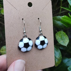 May include: A pair of silver dangle earrings with a black and white soccer ball charm. The earrings are on a brown card.