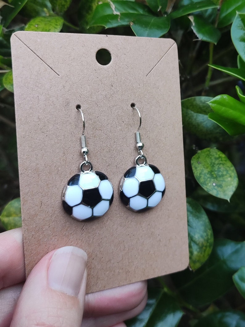 May include: A pair of silver dangle earrings with a black and white soccer ball charm. The earrings are on a brown card.
