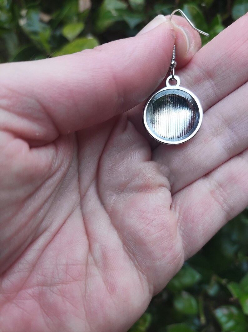 May include: Silver earring with a round, textured pendant. The pendant has a smooth, shiny surface and a slightly raised edge.