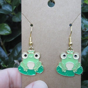 May include: A pair of gold dangle earrings featuring a green frog sitting on a green lily pad. The frog has black eyes and a white belly.