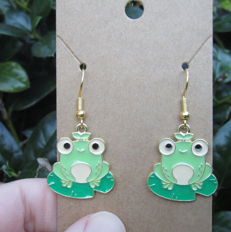 May include: A pair of gold dangle earrings featuring a green frog sitting on a green lily pad. The frog has black eyes and a white belly.