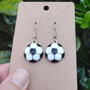 May include: A pair of silver earrings with black and white soccer ball charms. The earrings are displayed on a brown card.