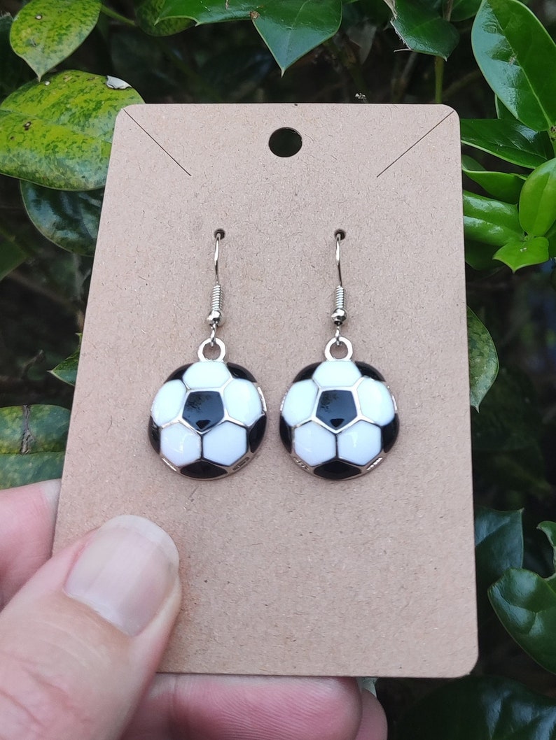 May include: A pair of silver earrings with black and white soccer ball charms. The earrings are displayed on a brown card.