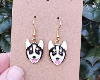 May include: A pair of gold-colored dangle earrings featuring a husky dog design. The earrings have a white face with black markings, blue eyes, and a pink tongue. The earrings are displayed on a brown card.