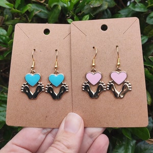 May include: Two pairs of gold earrings with a heart design. One pair has a light blue heart and the other pair has a pink heart. The hearts are held by two small feet.