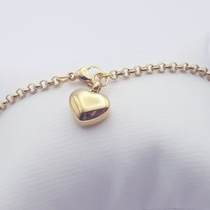 Könnte beinhalten: Goldfarbenes Armband mit herzförmigem Anhänger. Der Anhänger ist ein massives, poliertes Herz, das an einer zarten Kette hängt. Das Armband hat einen Karabinerverschluss und wird auf einer weißen Oberfläche präsentiert.