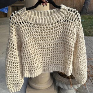 May include: A cream-coloured, open-weave crochet jumper with long sleeves and a crew neck. The jumper is held up on a black hanger. The crochet pattern creates a textured, airy design. The garment is likely handmade and suitable for layering.