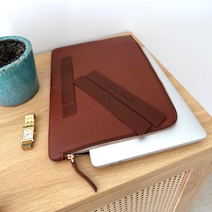 May include: A brown leather laptop sleeve with a zipper closure and a matching leather strap. The sleeve has a personalized leather strap with the name "John Smith" and initials "J.S." engraved on it. A gold watch and a blue ceramic pot are also visible.