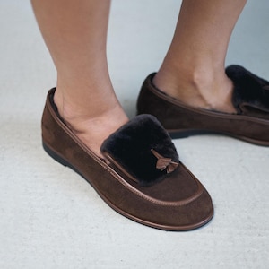 May include: Brown suede loafers with a dark fur accent. The shoes have a rounded toe and a small tassel detail. The soles are black. These comfortable shoes are designed for indoor wear.