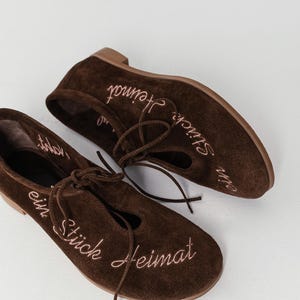 Pu&ograve; includere: Un paio di ballerine in camoscio marrone con un design stringato. Le scarpe hanno il testo ricamato "ein St&uuml;ck Heimat" in filo rosa. Le ballerine hanno la punta arrotondata e un tacco basso.