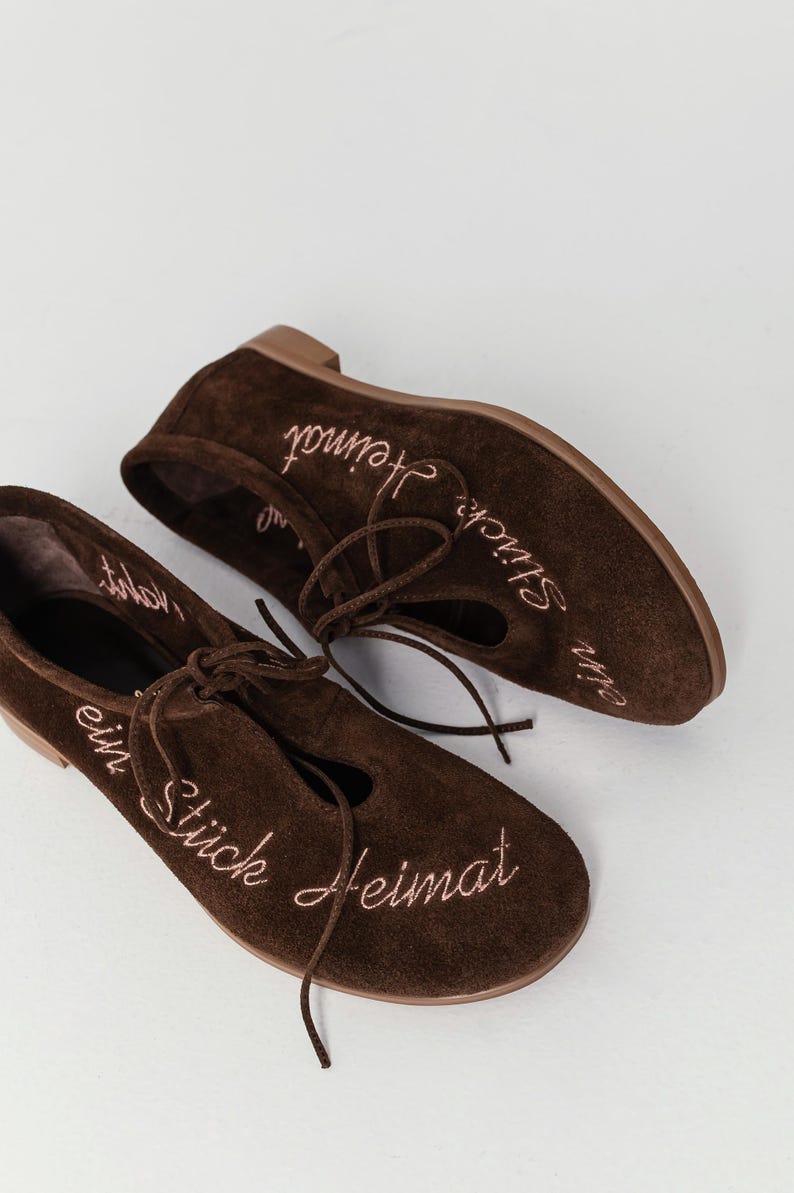 Pu&ograve; includere: Un paio di ballerine in camoscio marrone con un design stringato. Le scarpe hanno il testo ricamato "ein St&uuml;ck Heimat" in filo rosa. Le ballerine hanno la punta arrotondata e un tacco basso.