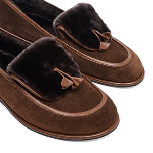 May include: A pair of brown suede loafers with dark brown fur accents and leather trim. Each shoe features a decorative tassel and a rounded toe. The shoes are designed for comfort and style.