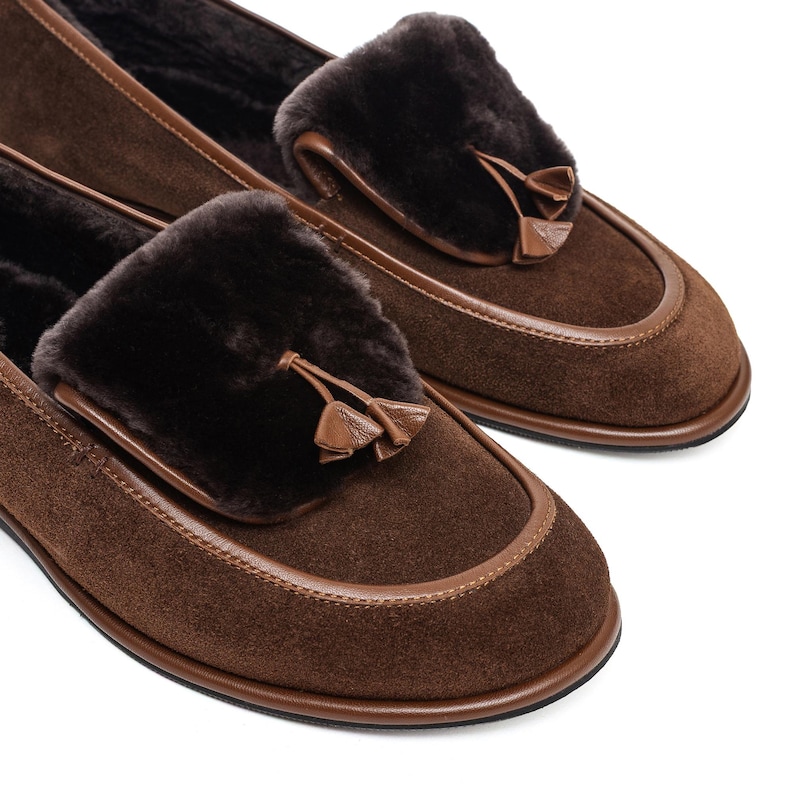 May include: A pair of brown suede loafers with dark brown fur accents and leather trim. Each shoe features a decorative tassel and a rounded toe. The shoes are designed for comfort and style.