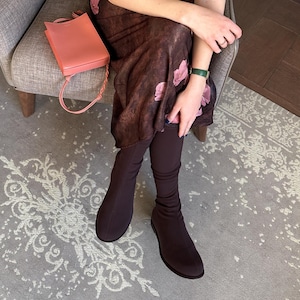 May include: A coral-coloured handbag with a braided strap is next to a person wearing a floral print dress and dark brown boots. The boots are a sleek, form-fitting style. A green bracelet is visible on the wrist.