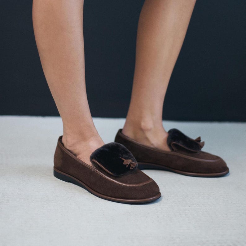 May include: Brown suede loafers with a dark fur accent. The shoes have a rounded toe and a small tassel detail. The soles are black. The shoes are on a light-coloured surface.