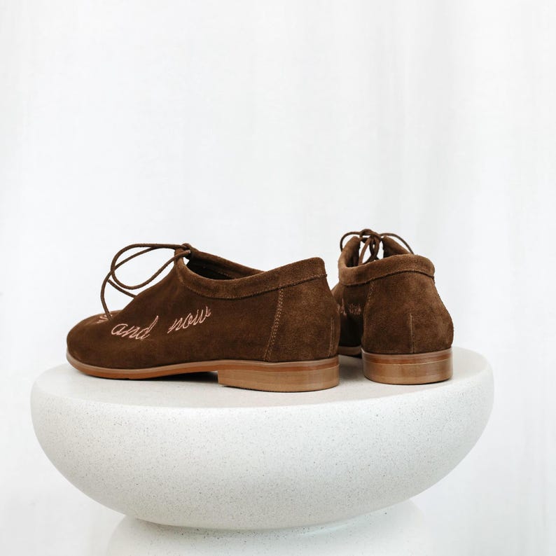 Pu&ograve; includere: Scarpe stringate in camoscio marrone con la scritta ricamata "and now". Le scarpe hanno un tacco basso e una suola marrone chiaro. Le scarpe sono esposte su un piedistallo bianco e arrotondato.