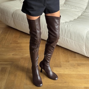 May include: Brown leather thigh-high boots with a pointed toe and a small heel. The boots have a zip on the side and are made of a smooth, shiny material. The boots are worn with black shorts.