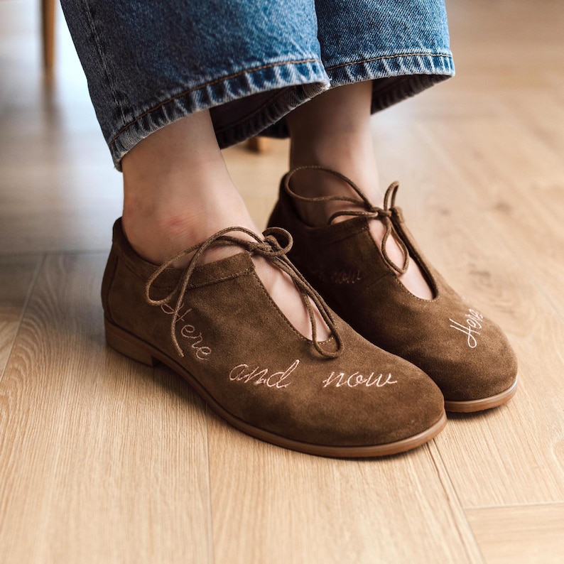 Pu&ograve; includere: Scarpe in camoscio marrone con design stringato. Le scarpe hanno le parole "here and now" ricamate sul lato. Le scarpe sono indossate con jeans blu.