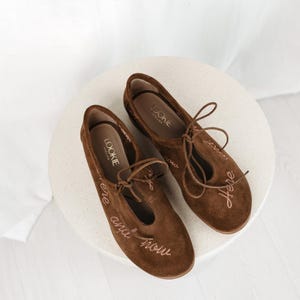 Pu&ograve; includere: Un paio di ballerine in camoscio marrone con le parole "here and now" ricamate sul lato. Le ballerine hanno un design a T e lacci. Il marchio "Lookie Atelier" &egrave; visibile sulla soletta.