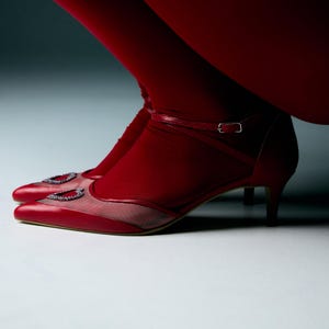 Puede incluir: Zapatos de tacón rojos de cuero con una correa y un adorno de piedras preciosas en la punta.