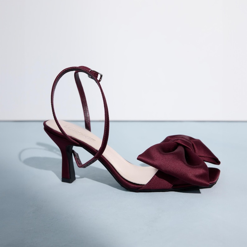 Op de afbeelding: Bordeauxkleurige satijnen sandalen met hak en een grote strik op de teen.
