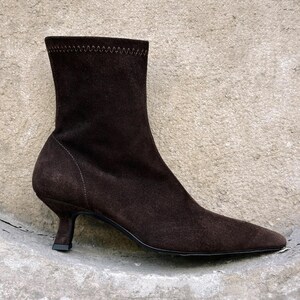 Könnte beinhalten: Braune Wildleder-Ankle-Boots mit spitzer Zehenpartie und kleinem, geschwungenem Absatz. Der Stiefel hat einen dehnbaren, gerippten Schaft und eine Nahtdetail. Der Absatz misst ca. 5 cm.