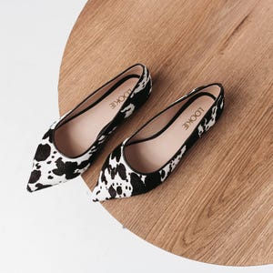May include: A pair of black and white cow print pointed toe flats. The shoes have a black trim and the brand name "LOOKIE" is printed on the insole.