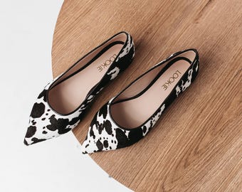 May include: A pair of black and white cow print pointed toe flats. The shoes have a black trim and the brand name "LOOKIE" is printed on the insole.
