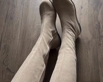 May include: A pair of knee-high boots in a light beige color with a smooth, matte finish. The boots feature a rounded toe, a vertical seam running up the front center, and a flat sole. The material appears soft and flexible, extending to the knee.