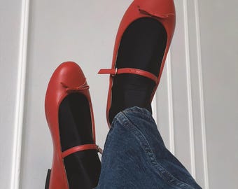 May include: A pair of red leather ballet flats with a small bow on the toe and a strap across the foot. The shoes are worn by a person wearing blue jeans.