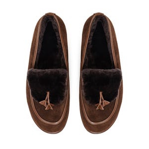 May include: A pair of brown suede loafers. They have a dark brown fur trim and a decorative tassel. The shoes have a rounded toe and a leather trim around the sole. The interior of the shoe is lined with a dark, soft material.
