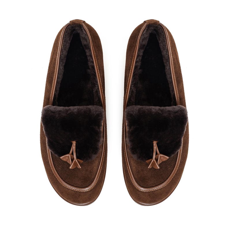 May include: A pair of brown suede loafers. They have a dark brown fur trim and a decorative tassel. The shoes have a rounded toe and a leather trim around the sole. The interior of the shoe is lined with a dark, soft material.